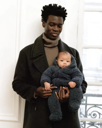 An African-American man in a coat holding a baby in a hooded snowsuit in front of a window, creating a heartwarming and stylish family moment.
