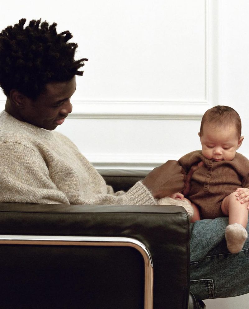 Young father sits in a modern chair holding his infant child on his lap, both facing the camera in an indoor setting against a white wall.