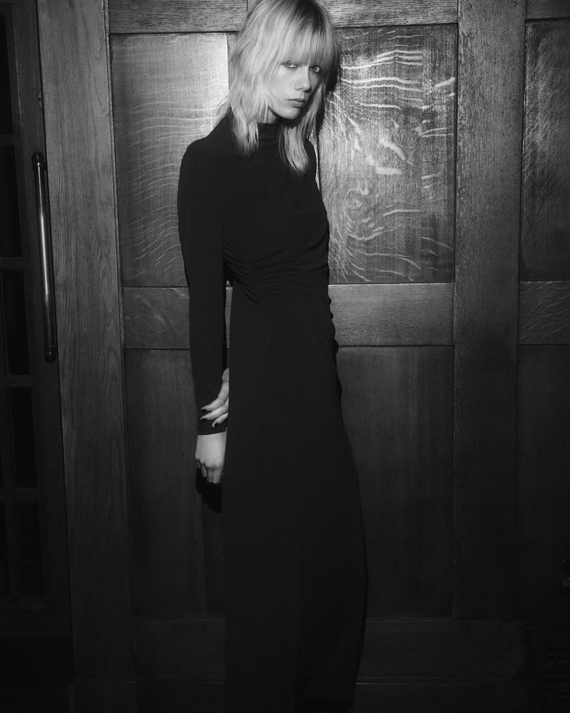 A young adult female model with blonde hair poses in a long black dress against a dark paneled door for a fashion shot in black and white.