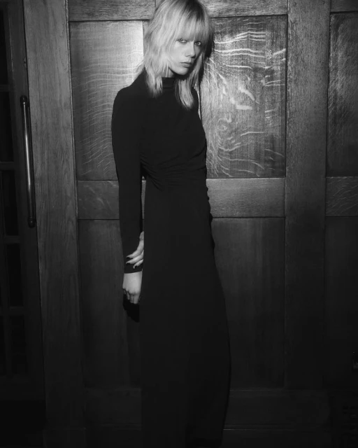 A young adult female model with blonde hair poses in a long black dress against a dark paneled door for a fashion shot in black and white.