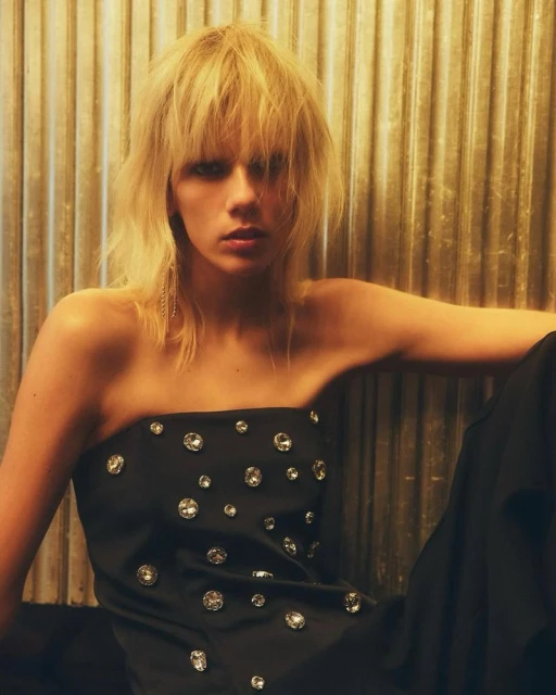 Blonde woman in jeweled black top poses against gold corrugated background in a fashion shot. Studio lighting, indoor environment.