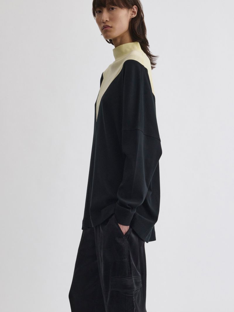 Fashion shot of a woman in a two-tone sweater and black pants against a plain background, showcasing modern style and minimalist design.