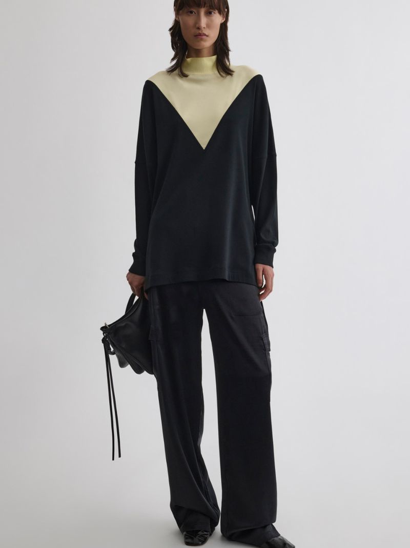 An adult woman is showcased indoors, wearing a stylish black and cream-colored long-sleeved top and loose black pants, holding a black bag.