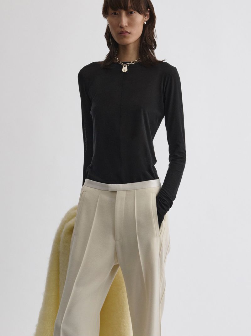 Woman in a black long-sleeve shirt and beige pleated pants poses in a studio with a pale yellow coat draped over her shoulder.