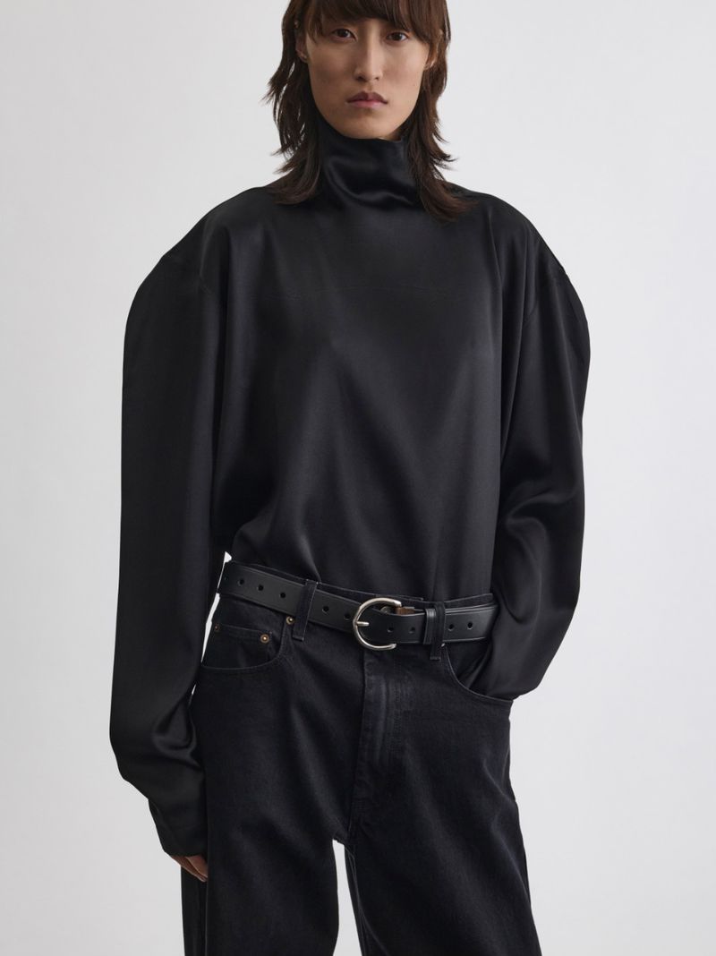 Model wearing a black silk turtleneck top and dark jeans with a leather belt against a white studio background
