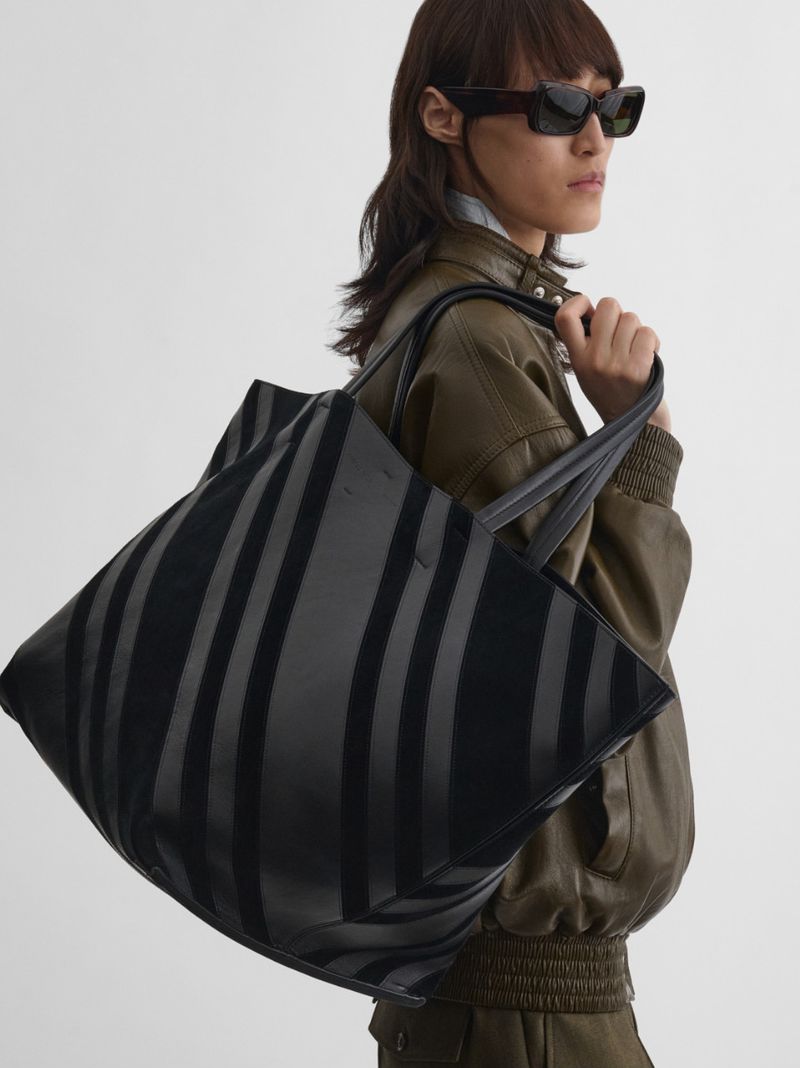 Woman in an olive green bomber jacket posing with a black striped tote bag in front of a neutral colored wall indoors.