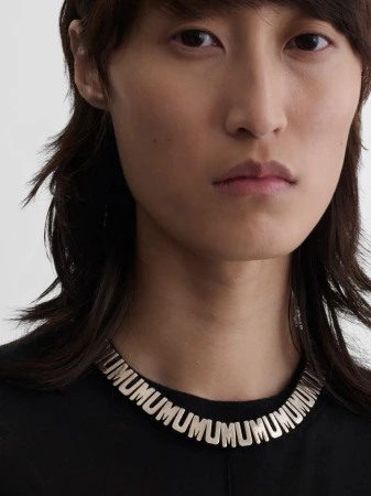 Close-up of an adult with dark hair and fair skin, wearing a black top and a silver necklace with repeating 'UMU' lettering.