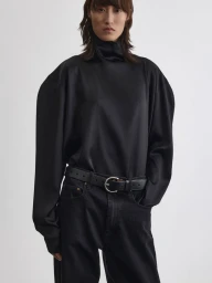 Model wearing a black silk turtleneck top and dark jeans with a leather belt against a white studio background