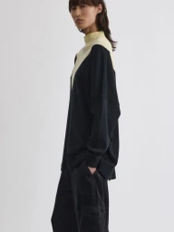 Model wearing a black and pale yellow mock neck top and dark trousers against a plain white background