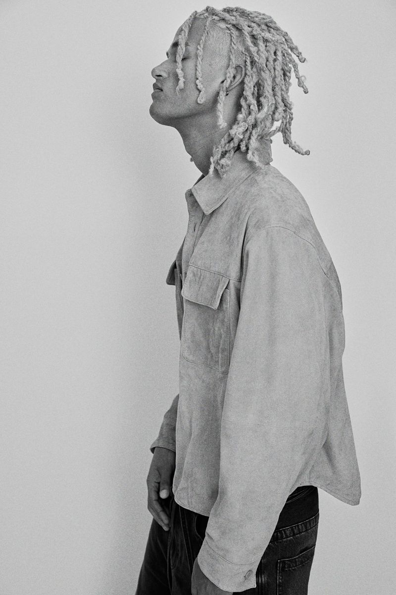 Profile portrait of a young man with blonde dreadlocks wearing a light suede shirt against a plain light background