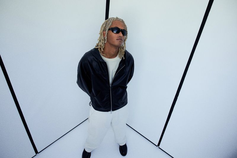 A young adult male with dreadlocks stands in a white room, wearing sunglasses, a black jacket, and white pants in an eye-level medium shot.