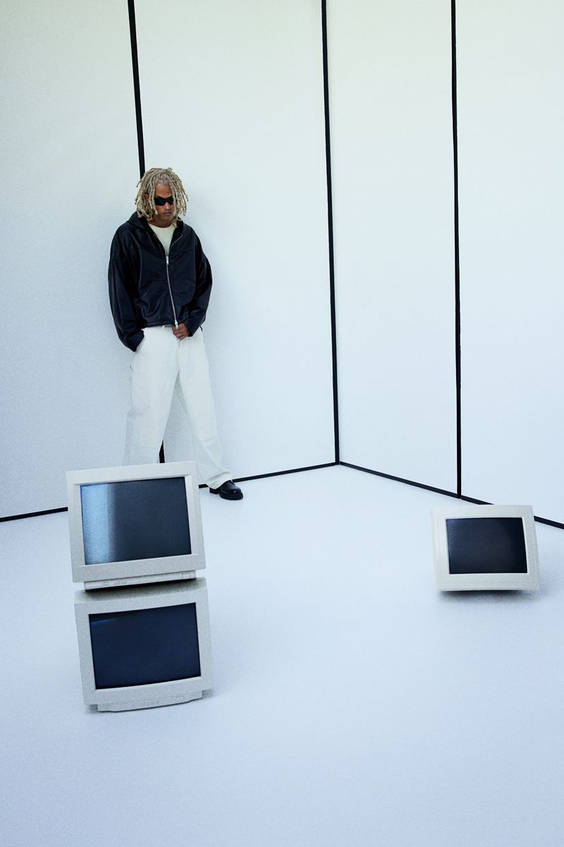 An adult with blonde hair stands in a studio with white walls and two vintage monitors with a third monitor in the background.