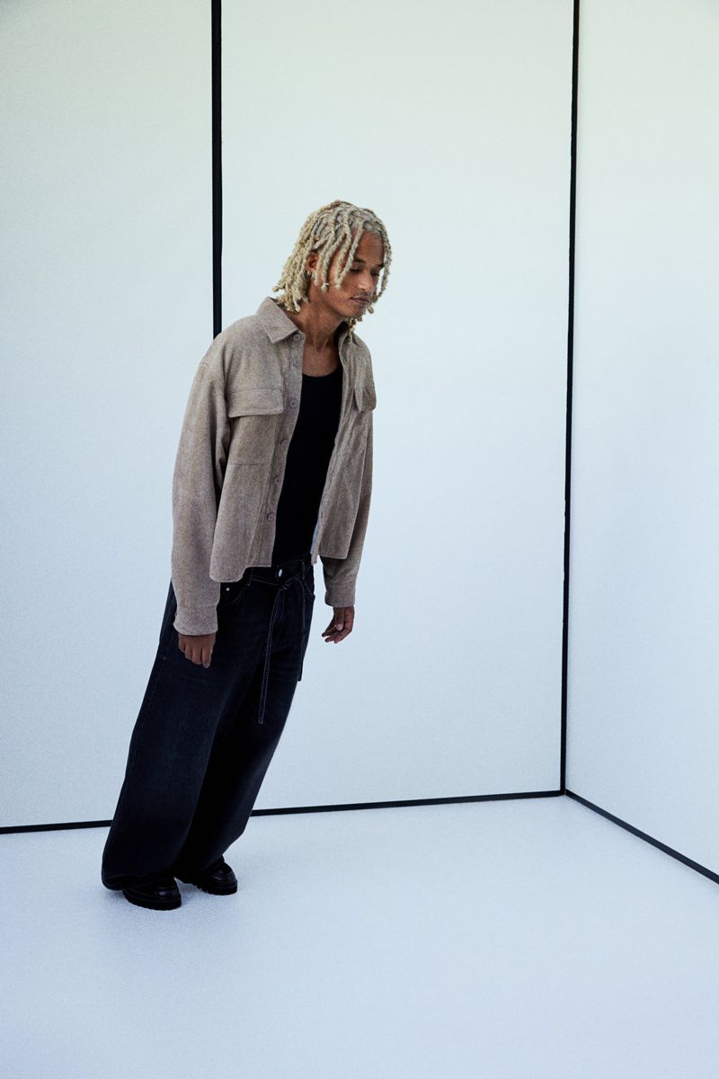 Young adult poses in front of corner studio panels wearing a beige collared shirt, black tank top, and dark jeans with long blonde dreadlocks.