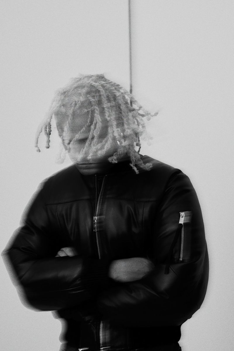 Monochrome portrait of adult with dreadlocks, wearing a black puffer jacket with arms crossed, standing in front of a white wall.