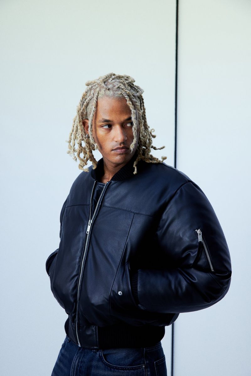 Portrait of a young adult male with blond dreadlocks wearing a black bomber jacket and blue jeans against a white background.