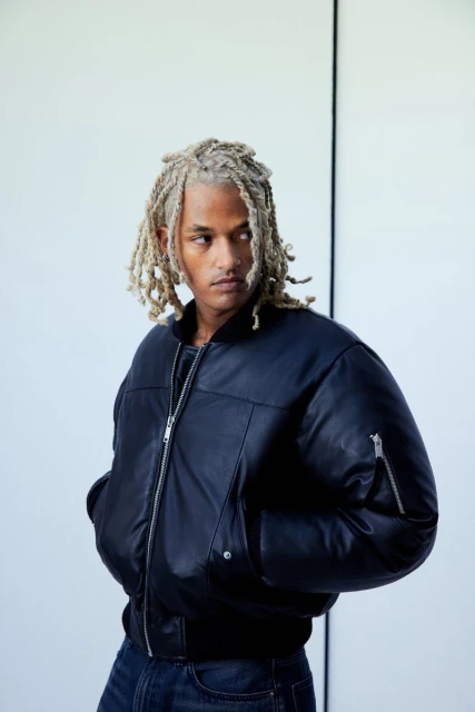 Portrait of a young adult male with blond dreadlocks wearing a black bomber jacket and blue jeans against a white background.