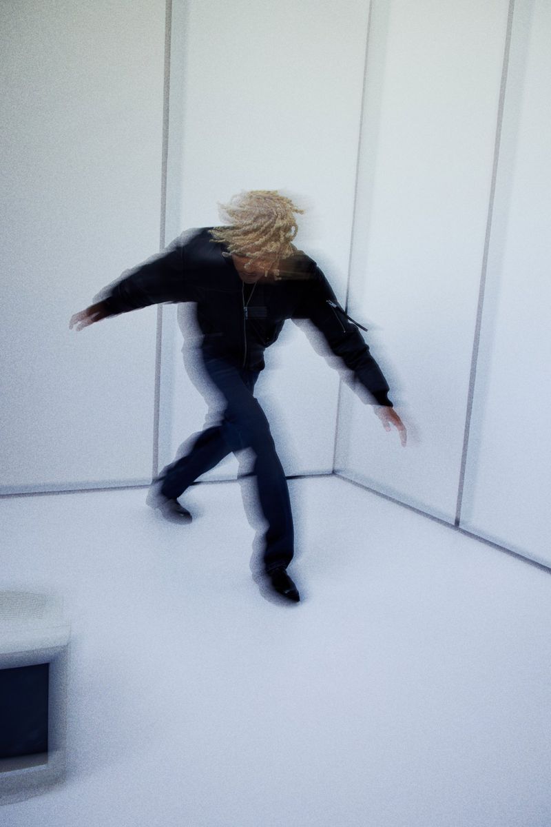 Motion-blurred shot of a person with blond dreadlocks in a black jacket and blue jeans moving in a white indoor space with neutral lighting.