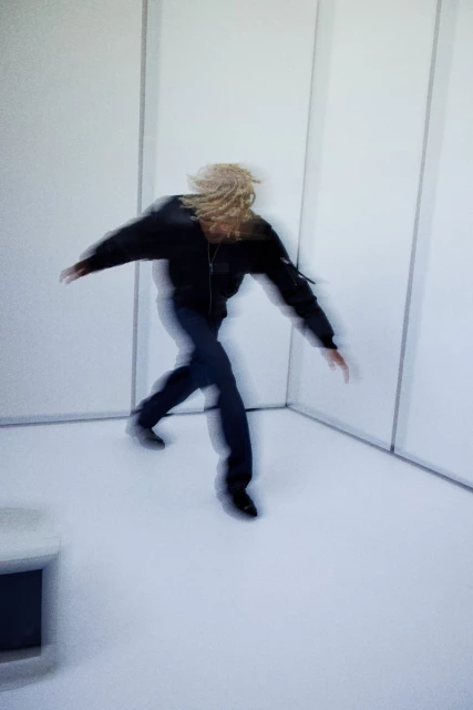 Motion-blurred shot of a person with blond dreadlocks in a black jacket and blue jeans moving in a white indoor space with neutral lighting.
