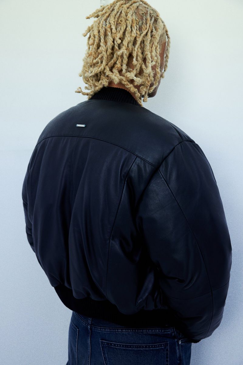 Adult human with blond dreadlocks wears a black leather bomber jacket and blue jeans against a white wall in a studio shot.