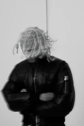 Man with braided blonde hair wearing a black leather jacket with arms crossed against a plain white background