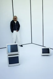 Model wearing a black jacket and white trousers standing in a white studio set with vintage computer monitors.