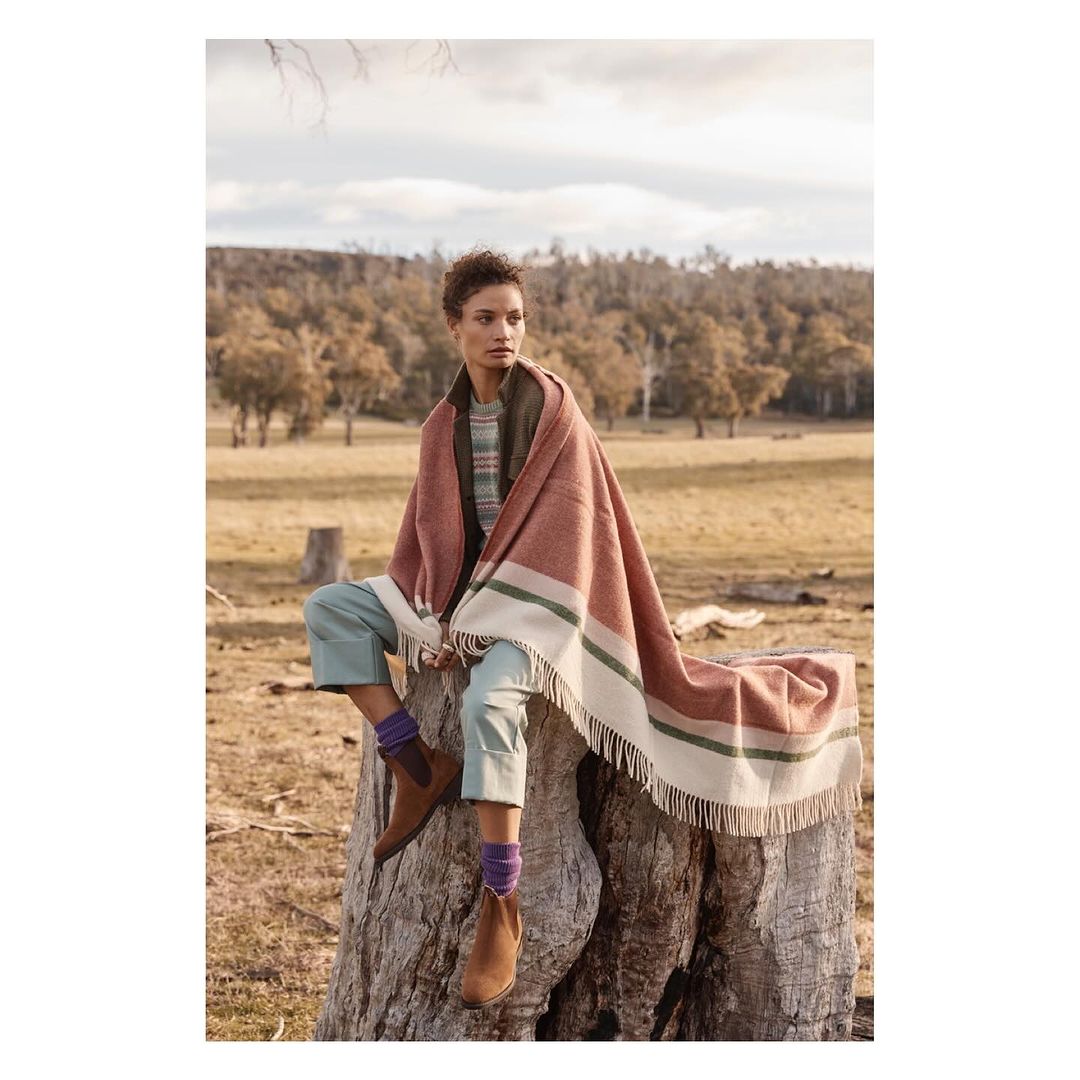Woman with short hair sits on tree stump wrapped in terracotta and cream striped fringed blanket in dry rural landscape setting.