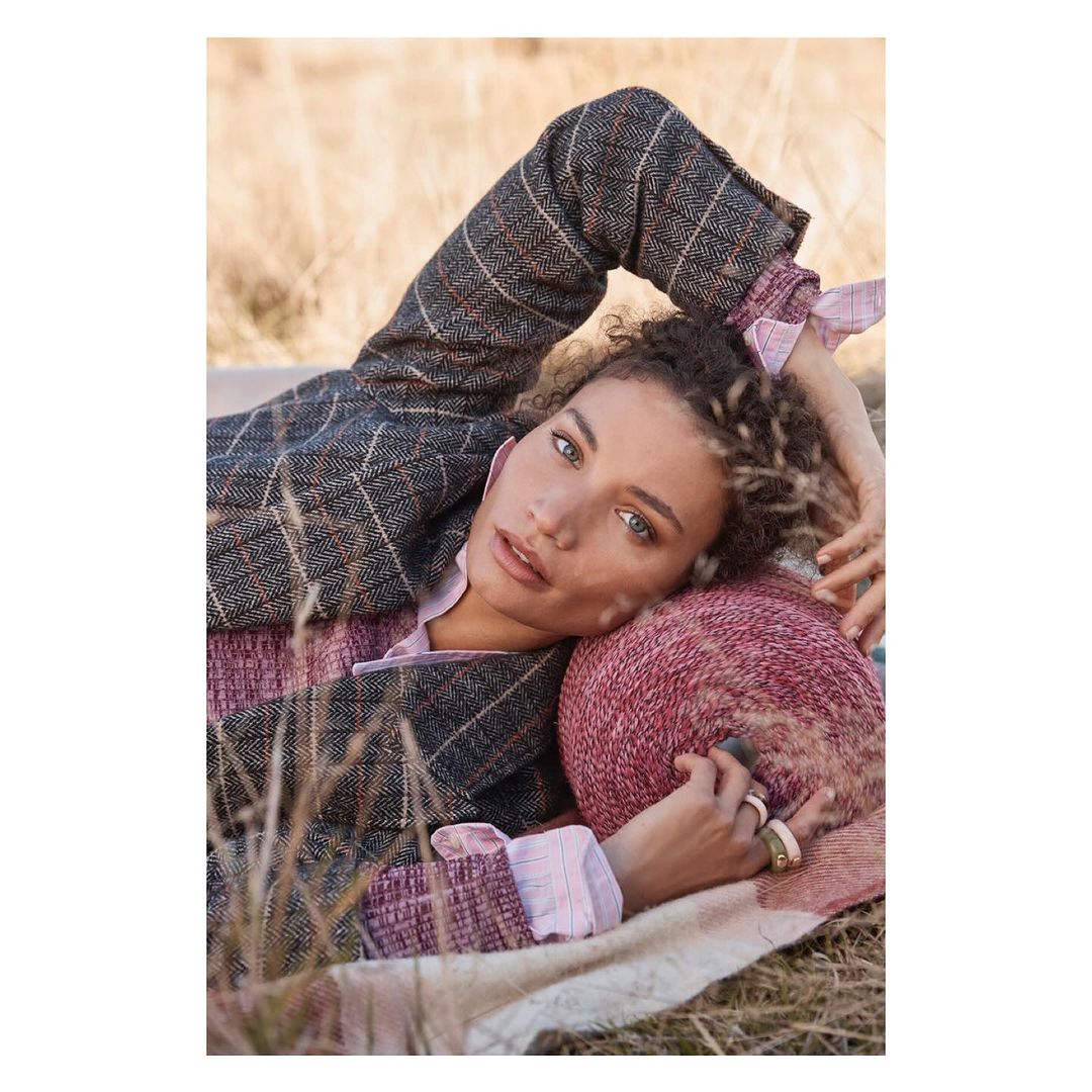Adult female model in a brown tweed jacket and pink shirt lying in a grassy field with a pink knit pillow and a cream blanket.