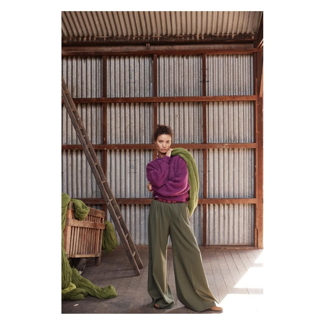 A woman in a purple sweater and olive pants stands inside a corrugated metal barn, a green shawl over her shoulder, with a ladder nearby.