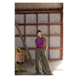Model wearing a purple sweater and olive green trousers posing inside a rustic shed with corrugated metal walls