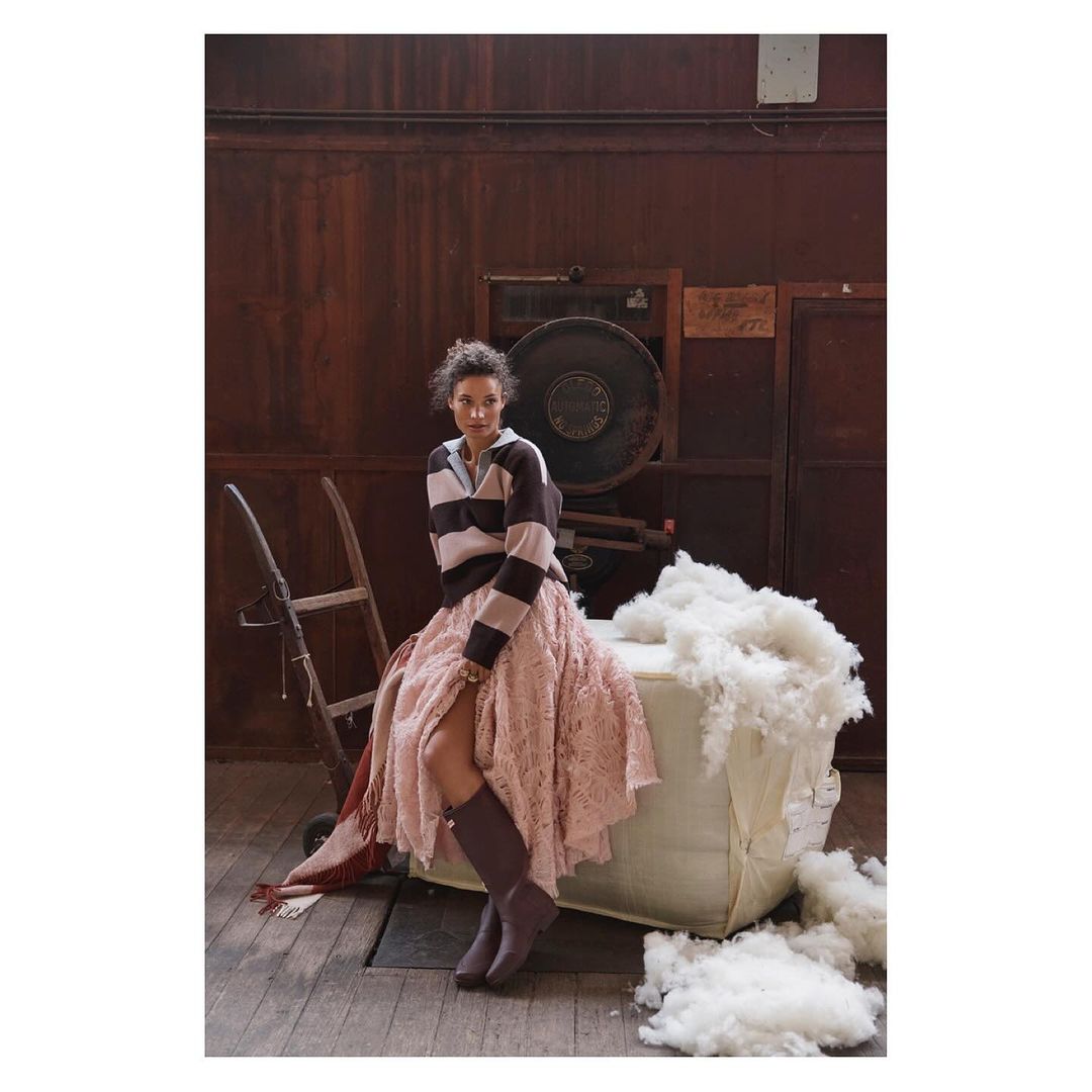 A woman in a striped sweater and pink skirt sits on a white bale in a rustic interior with a vintage machine and wooden floors.