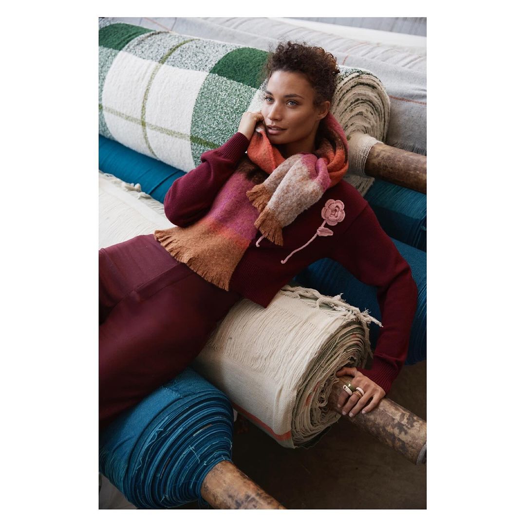 A woman in a maroon sweater and pants, with a pink and orange scarf, poses while reclining on rolls of colorful fabric indoors.