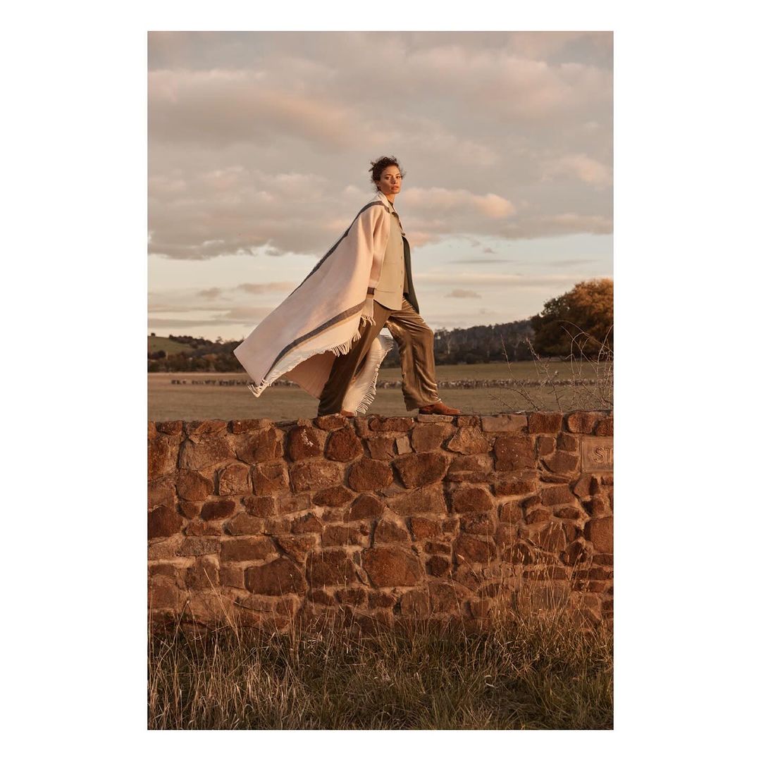 Woman in tailored suit and cape posing on stone wall in open field with cloudy sky. Fashion editorial photography in natural outdoor light.