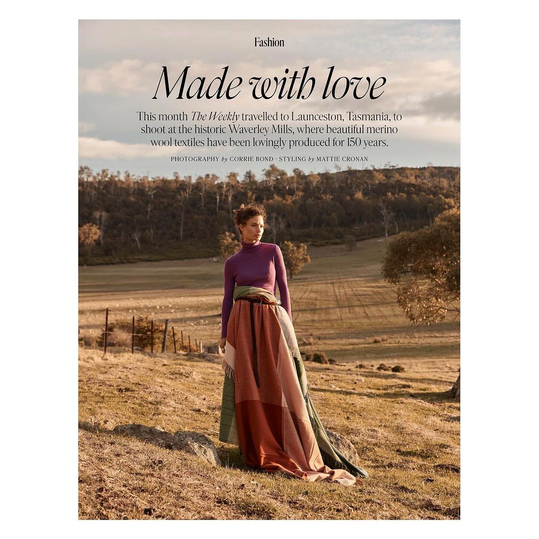 Woman in a stylish long skirt stands in a rural field with rolling hills and trees, modeling for a fashion editorial shoot.