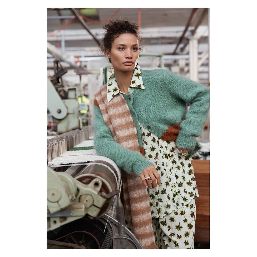Fashionable adult female in a patterned dress and cardigan posing near industrial machinery in a bright, indoor setting with natural light.