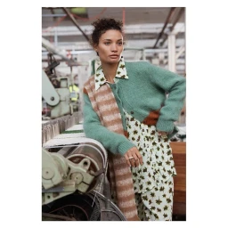 Woman leaning on a textile machine wearing a green cardigan, patterned shirt, and striped scarf inside a factory.