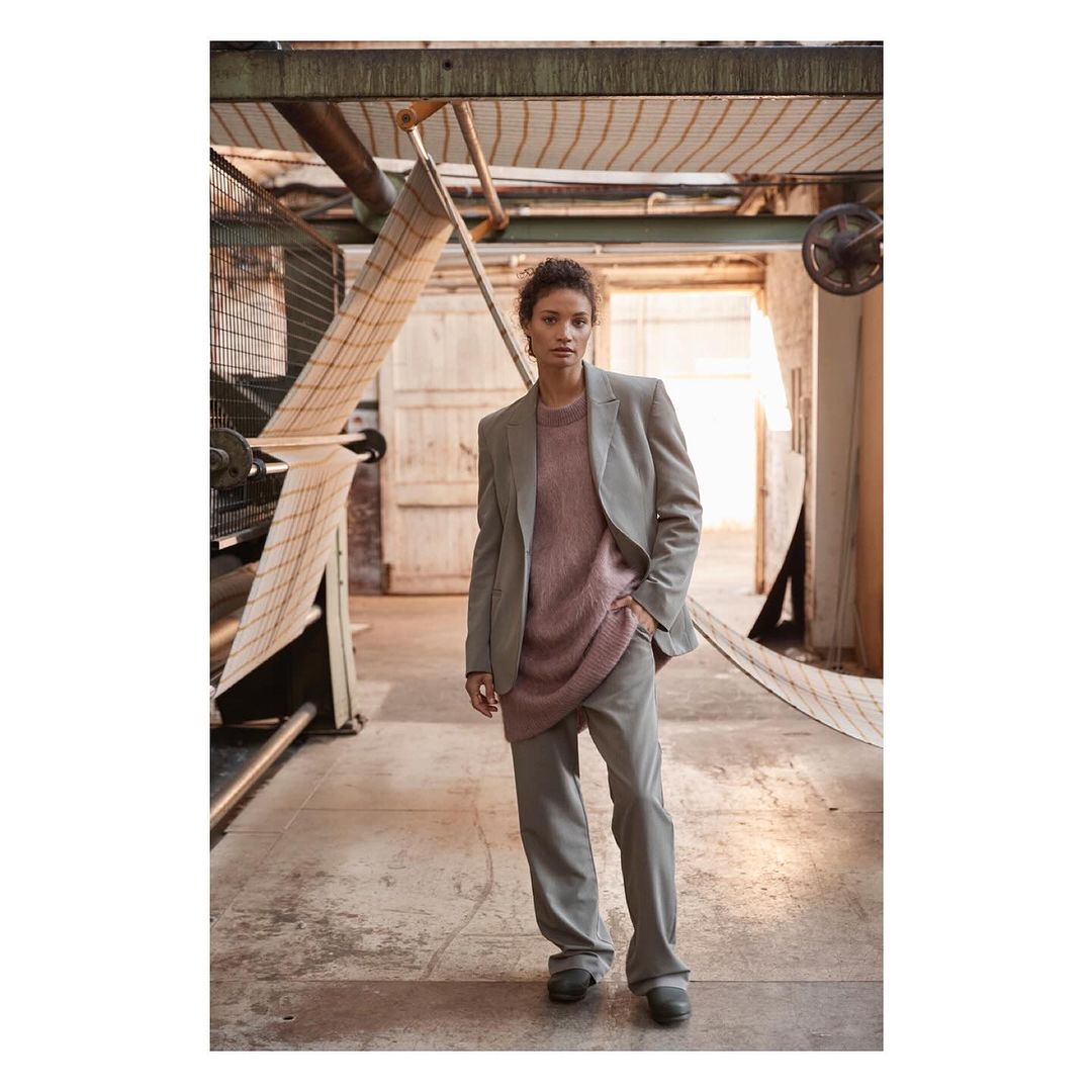 Stylish woman in grey suit and pink sweater posing indoors with natural light and industrial background elements, fashion editorial shot.