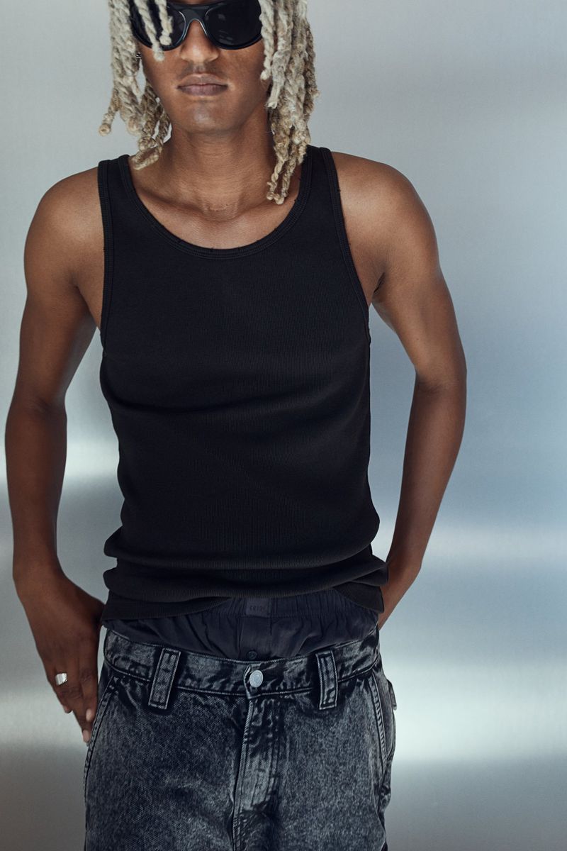 Portrait of a man in black tank top and jeans wearing black sunglasses against a silver background in a studio environment.