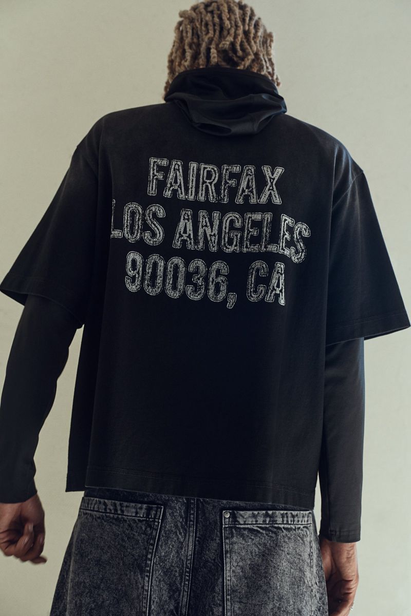Person with blond dreadlocks wears a black t-shirt with 'Fairfax Los Angeles 90036, CA' printed on the back, standing indoors.