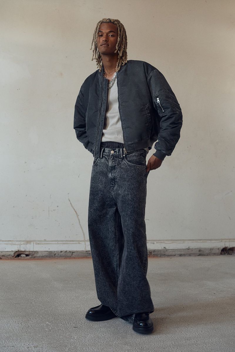 Full-length shot of adult male with blond dreadlocks in gray t-shirt, black bomber jacket, wide-leg denim jeans, and black shoes in studio.
