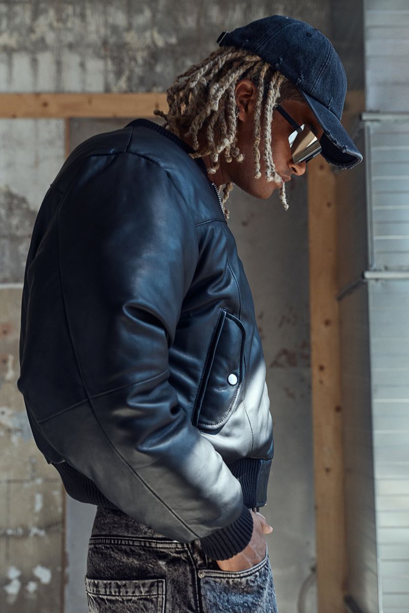 Man in black leather jacket and cap poses against a neutral indoor background with sunlight, showcasing fashion and style.