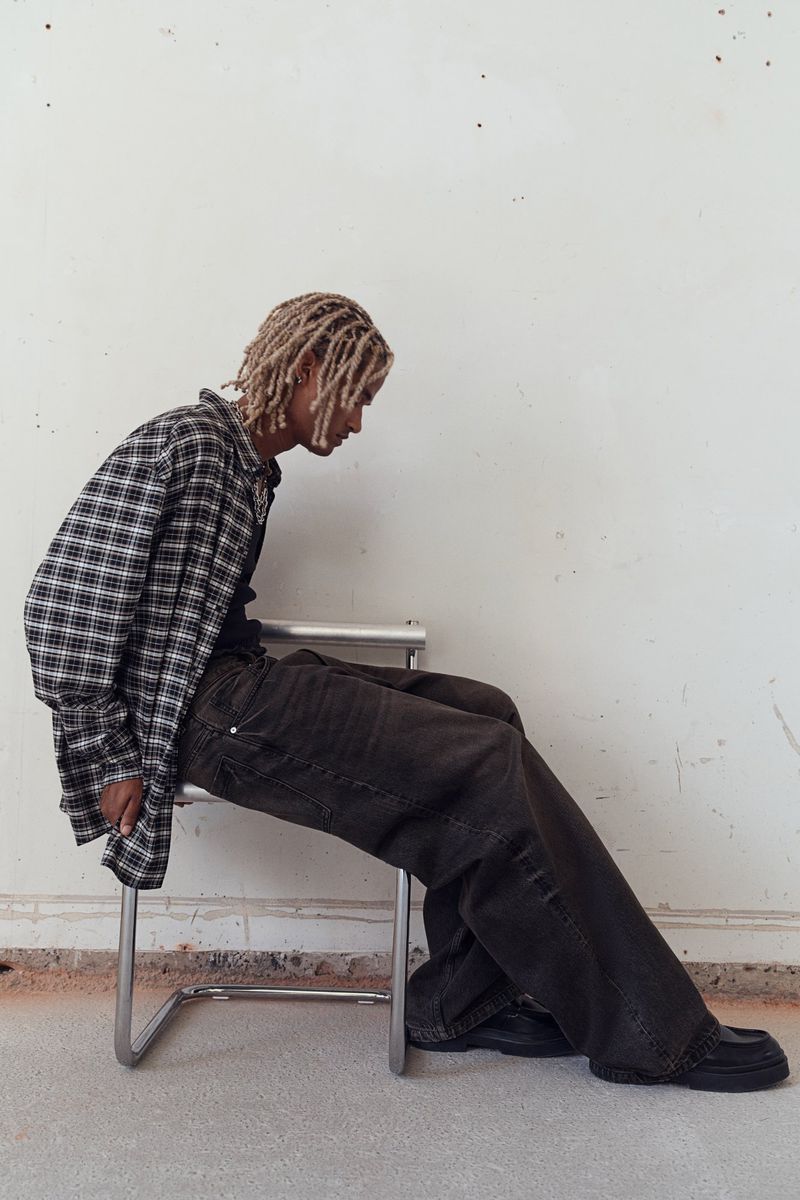 Person with dreadlocks wearing a flannel shirt and dark denim pants sitting on a metal chair against a white wall indoors.
