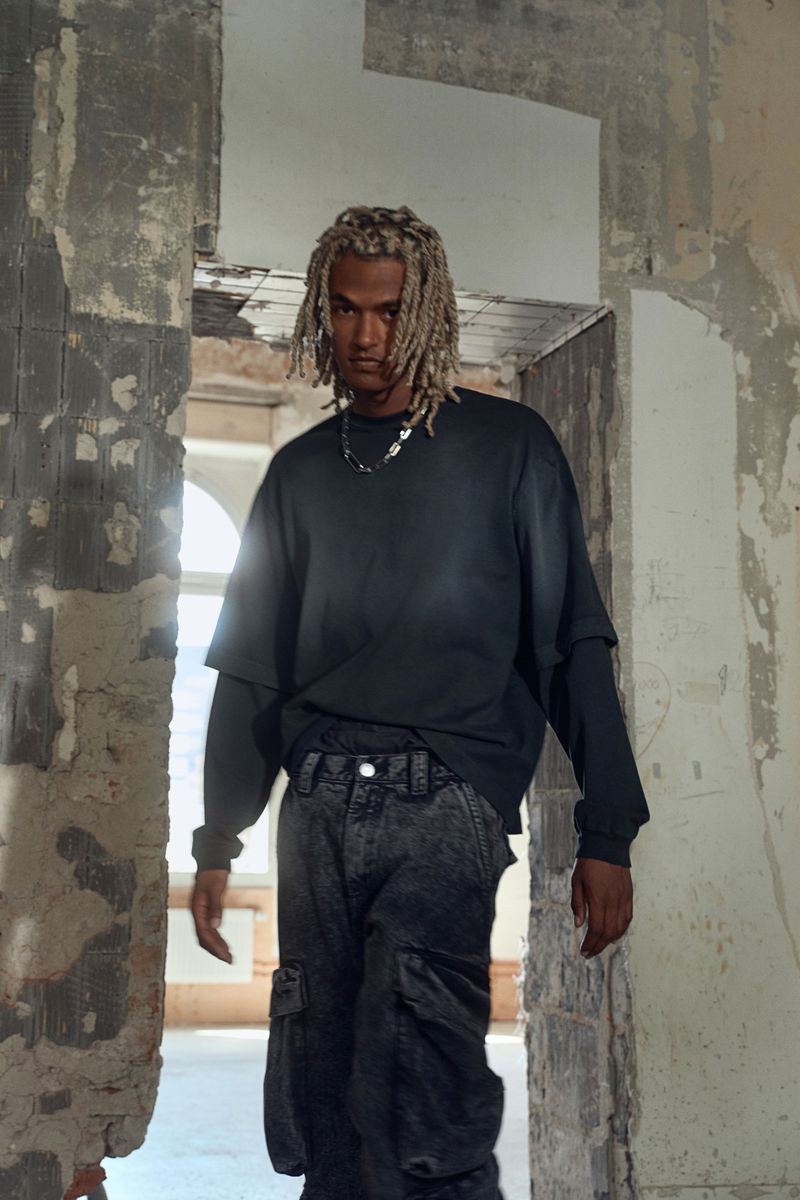 Man in oversized black sweatshirt and cargo pants posing in ruined doorway with neutral facial expression looking at camera indoors.