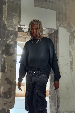 Man in oversized black sweatshirt and cargo pants posing in ruined doorway with neutral facial expression looking at camera indoors.