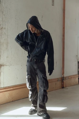 Full-length portrait of one adult male with blond dreadlocks wearing black hooded jacket, grey jeans, and black sneakers in an indoor setting.