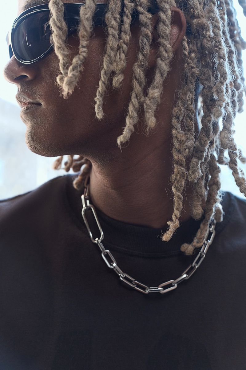 Close up of a person with dreadlocks wearing sunglasses and a silver chain necklace against a bright background.