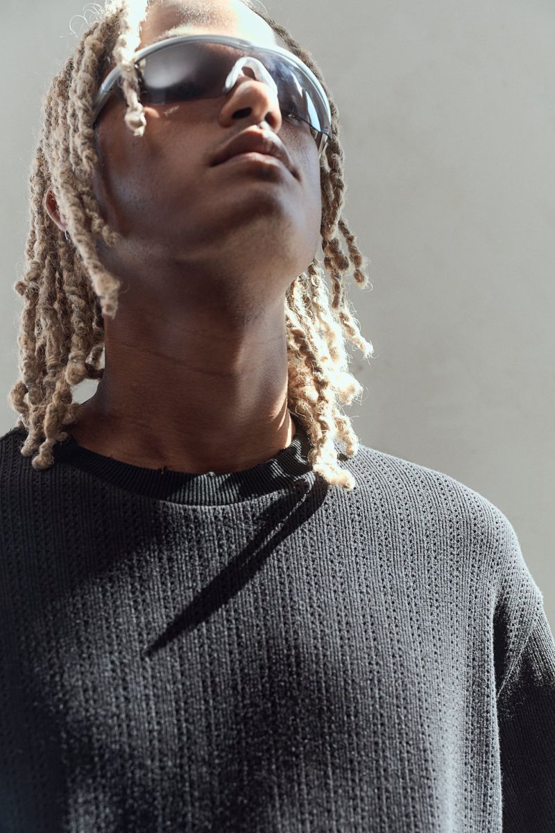 Low-angle portrait of a black male with blonde dreadlocks wearing a black shirt and sunglasses looking up against a grey backdrop.