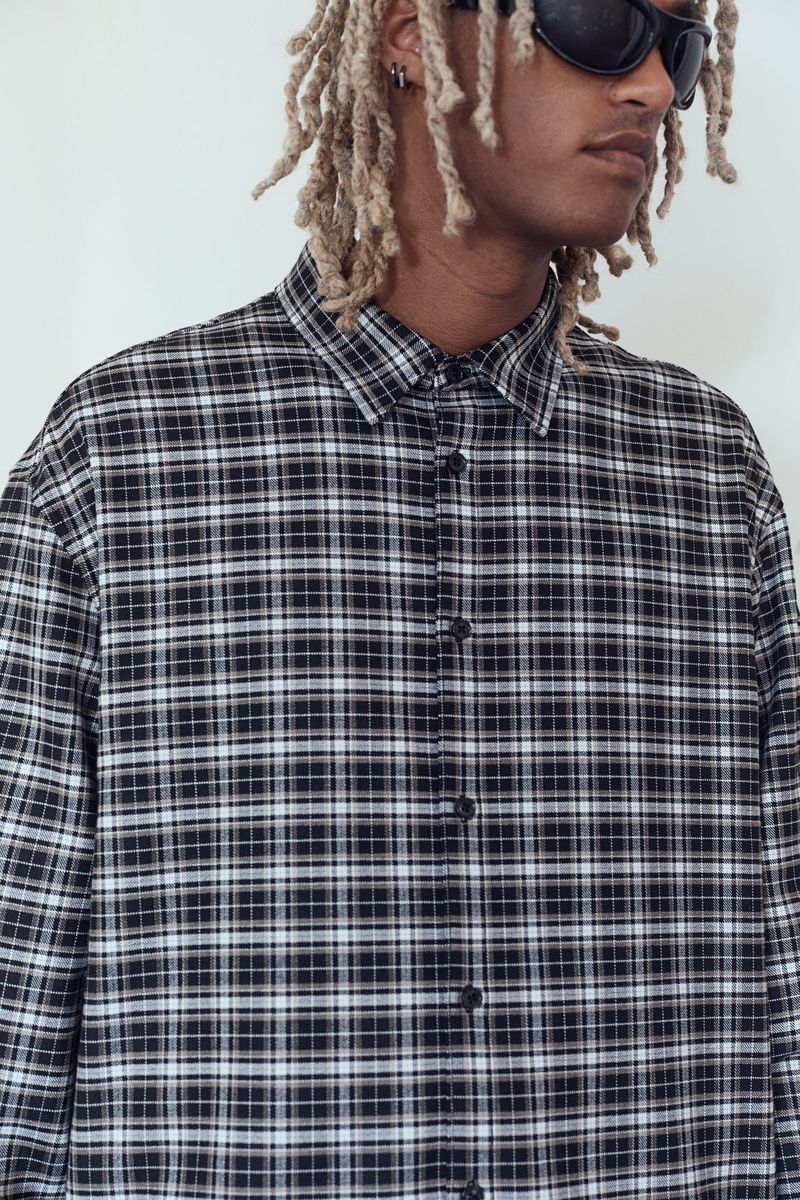 Adult male poses indoors wearing a black and white plaid collared button-down shirt, sunglasses, and blonde dreadlocks against a white wall.