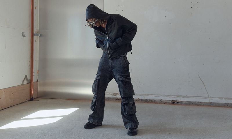 Fashionable person in an oversized black jacket and baggy cargo pants poses in a minimalist studio with concrete floors and white walls.