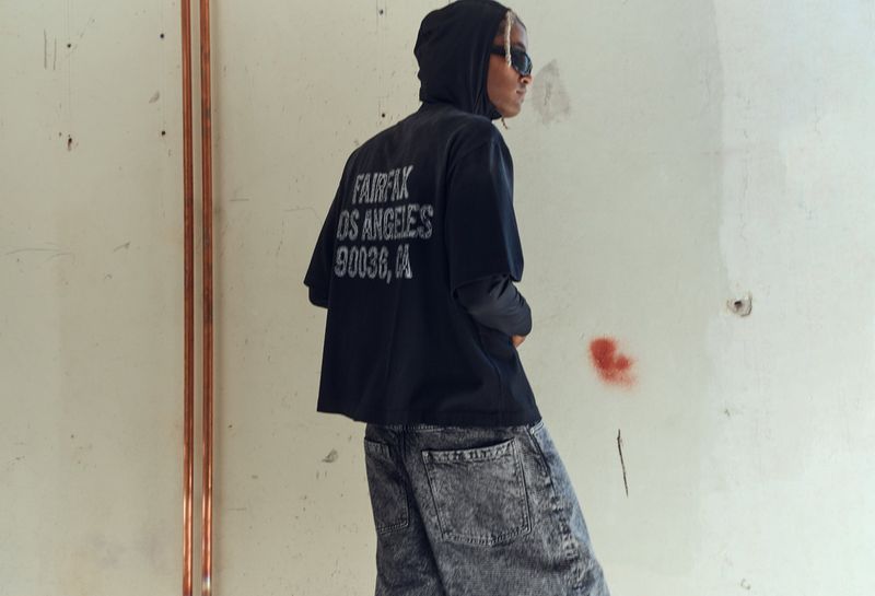 Young adult poses in black graphic t-shirt and grey jeans against an industrial background, wearing sunglasses and a hoodie.