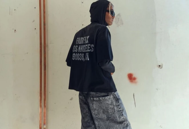 Young adult poses in black graphic t-shirt and grey jeans against an industrial background, wearing sunglasses and a hoodie.
